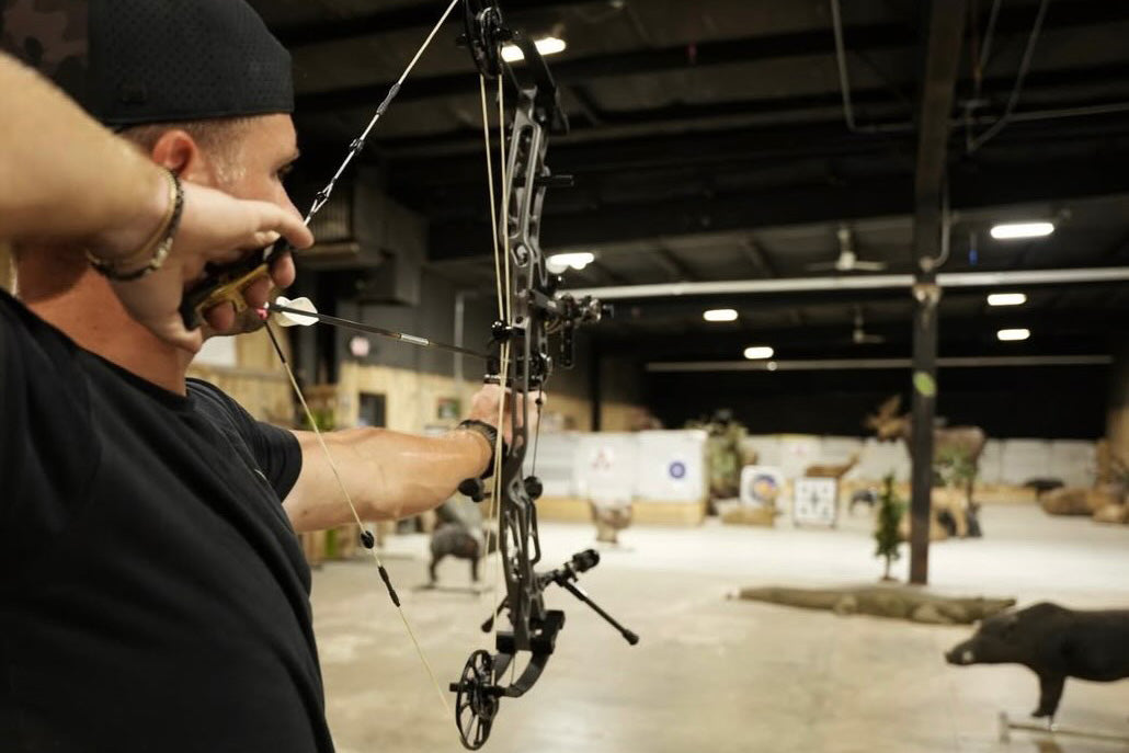Man preparing a bow and arrow indoors with a large indoor shooting range in the background.