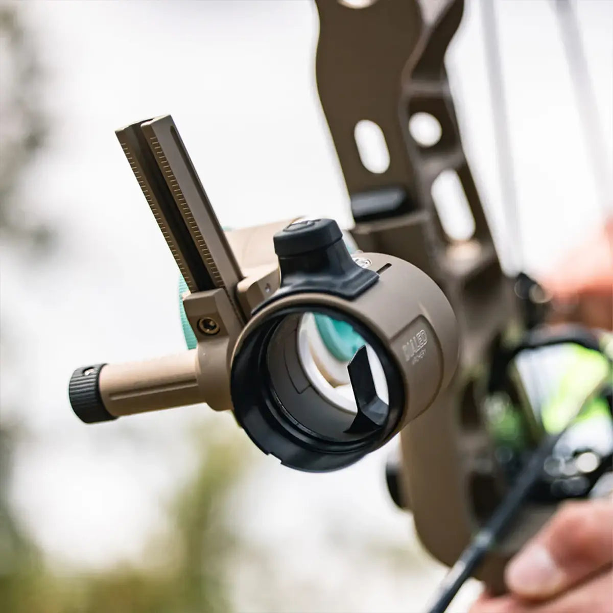 Tan-colored archery dialed sight lens with black adjustment knobs and clear glass lens mounted on a bow.