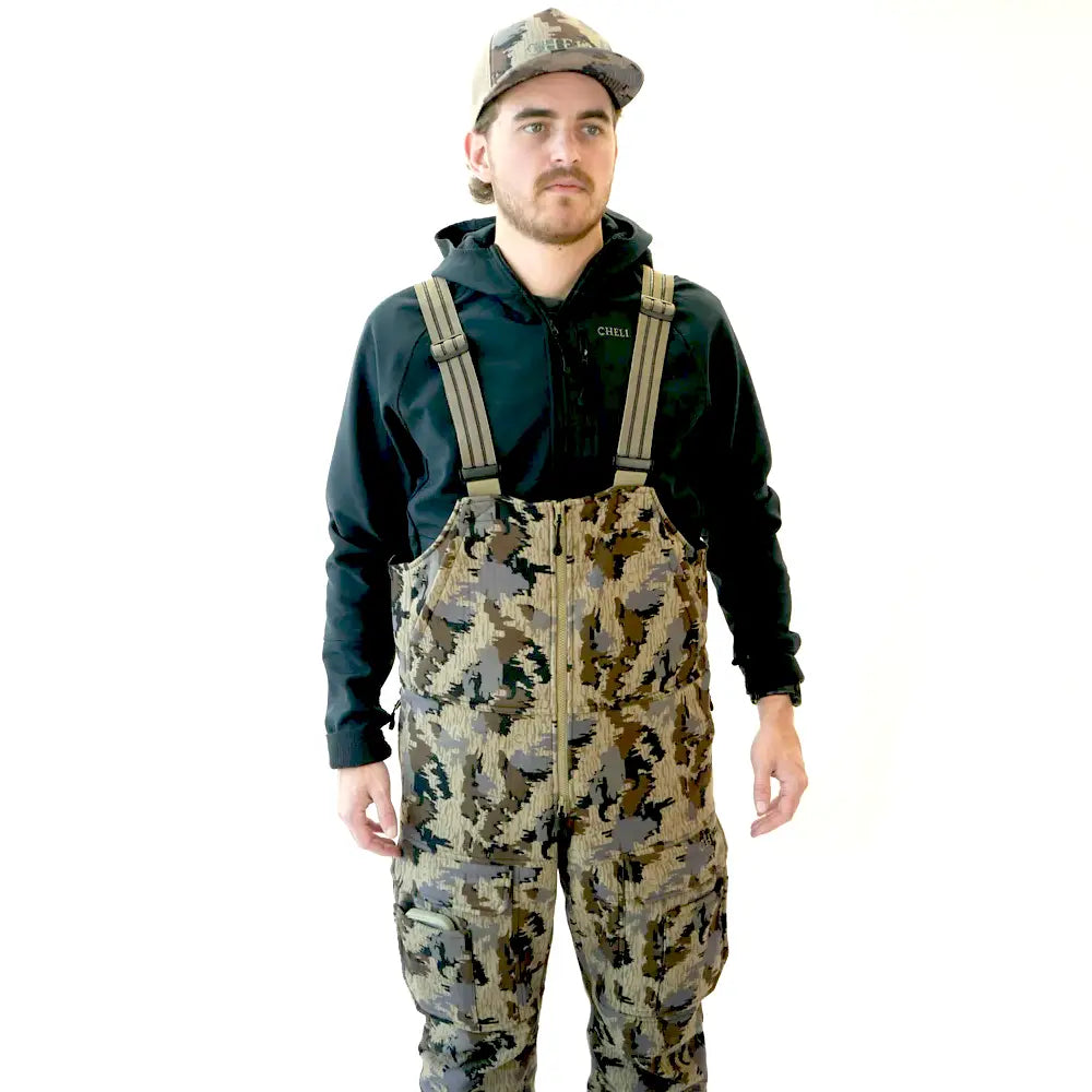 A man wearing camouflage-patterned waders over a black hoodie and a matching camouflage cap, standing against a plain white background.