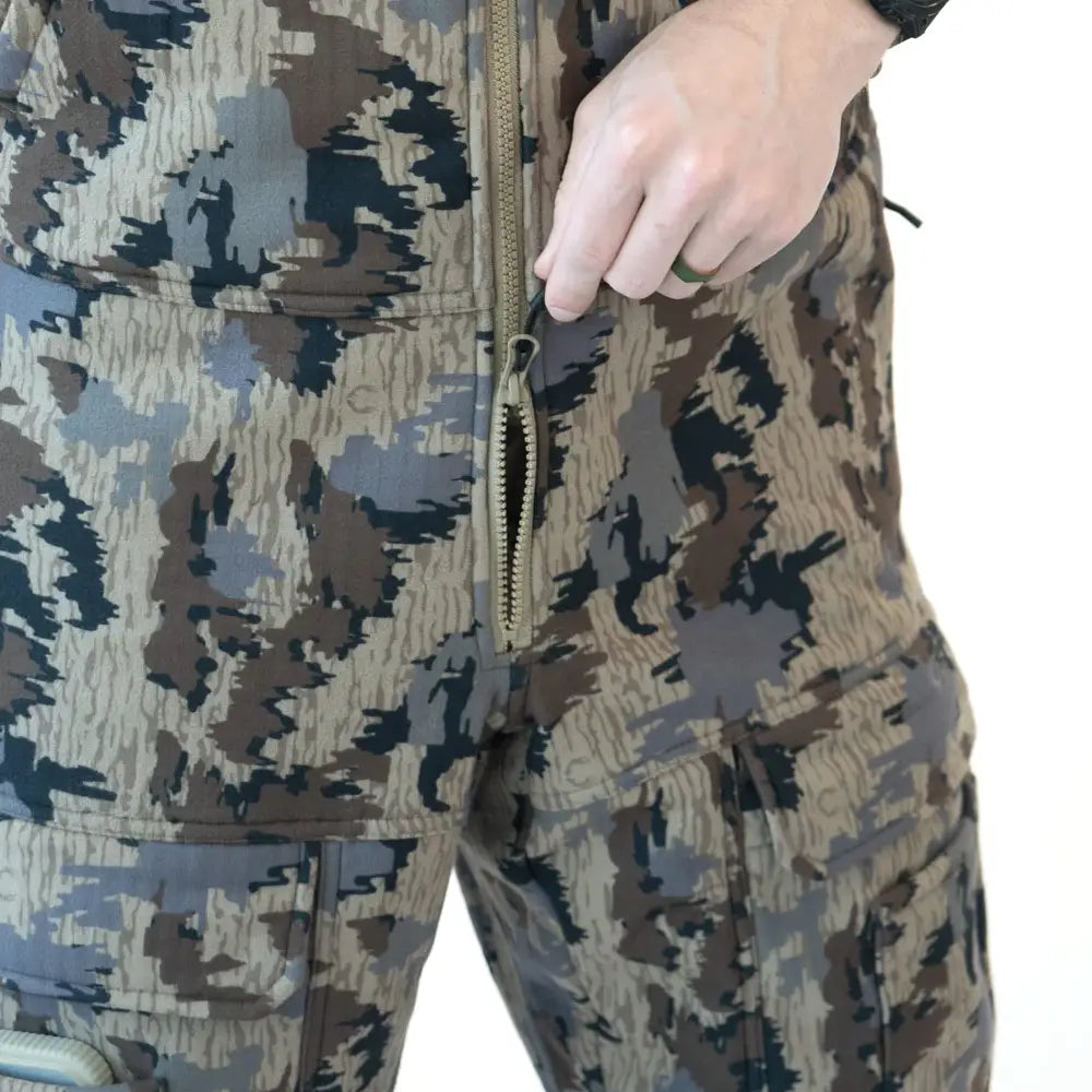 Close-up of a person wearing camouflage pants, showing a hand pulling a zipper on the front of the garment. The pants feature a multi-tonal camo pattern in beige, brown, gray, and black.