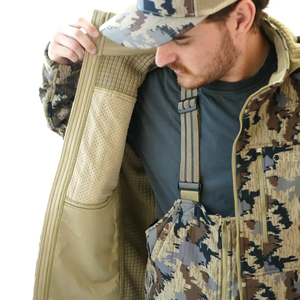 A man wearing a camouflage hunting jacket and matching camo cap, opening the jacket to show the tan interior lining.