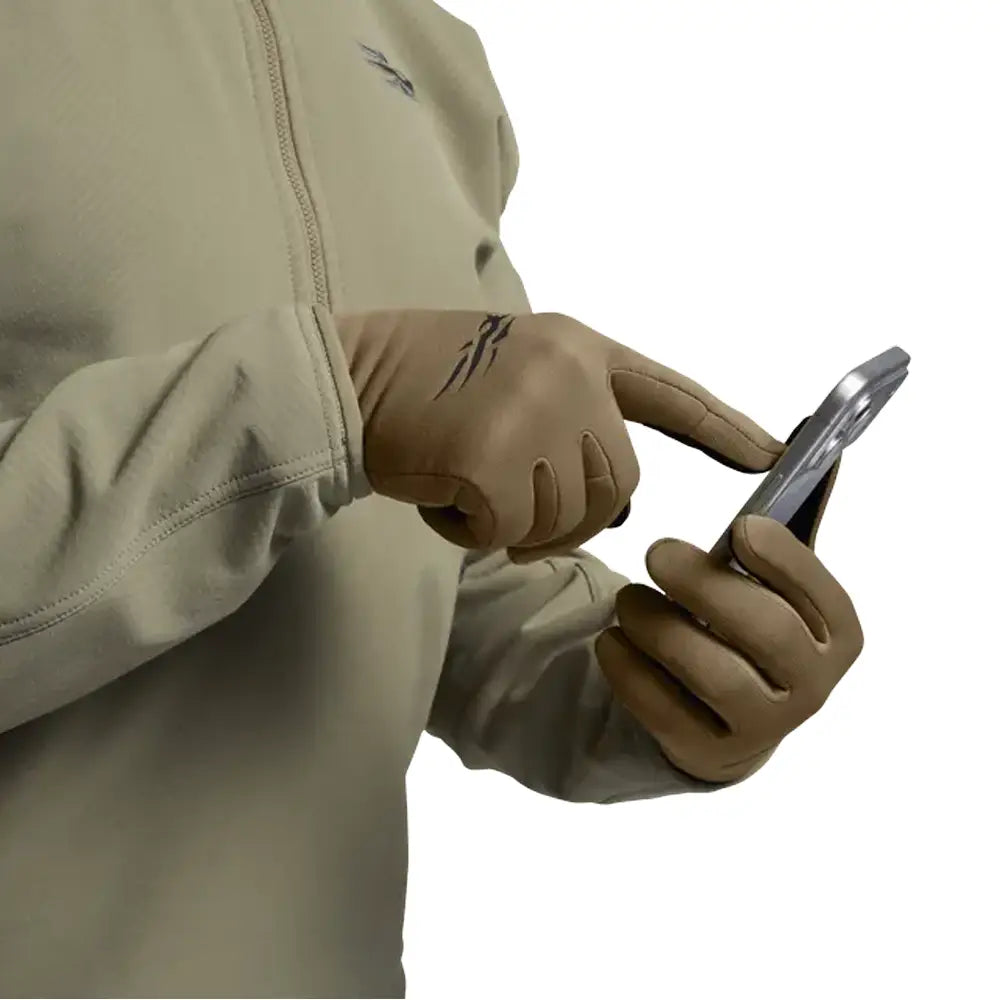 A close-up of a person wearing tan tactical gloves, with one hand touching a smartphone screen while the other holds the phone. The person is also wearing a light beige zip-up jacket against a plain white background.